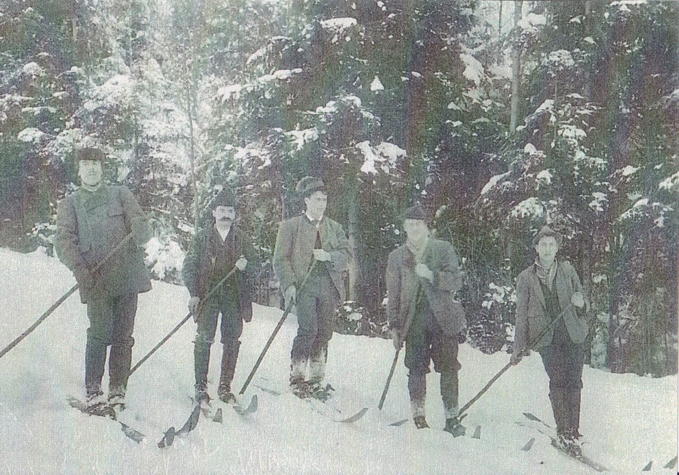 Fünf Männer in Lodenjacken stehen mit langen Holzski und je einem Stock im verschneiten Wald.
