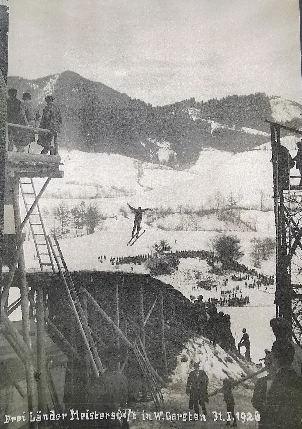 Ein Skispringer fliegt mit ausgebreiteten Armen über eine hölzerne Schanze, am Gegenhang stehen Zuschauer in langen Reihen.