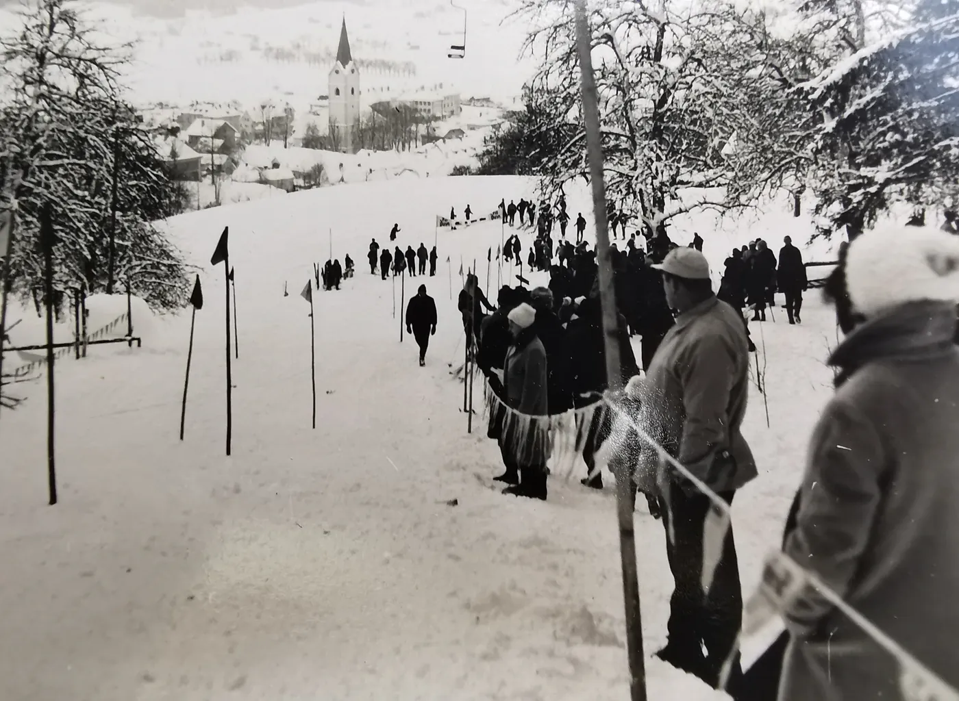 Ein Torlauf mit Holzstangen und Fähnchen auf einem verschneiten Hang, am Rand Zuschauer, im Tal die Pfarrkirche, darüber ein Sessel des Lifts.
