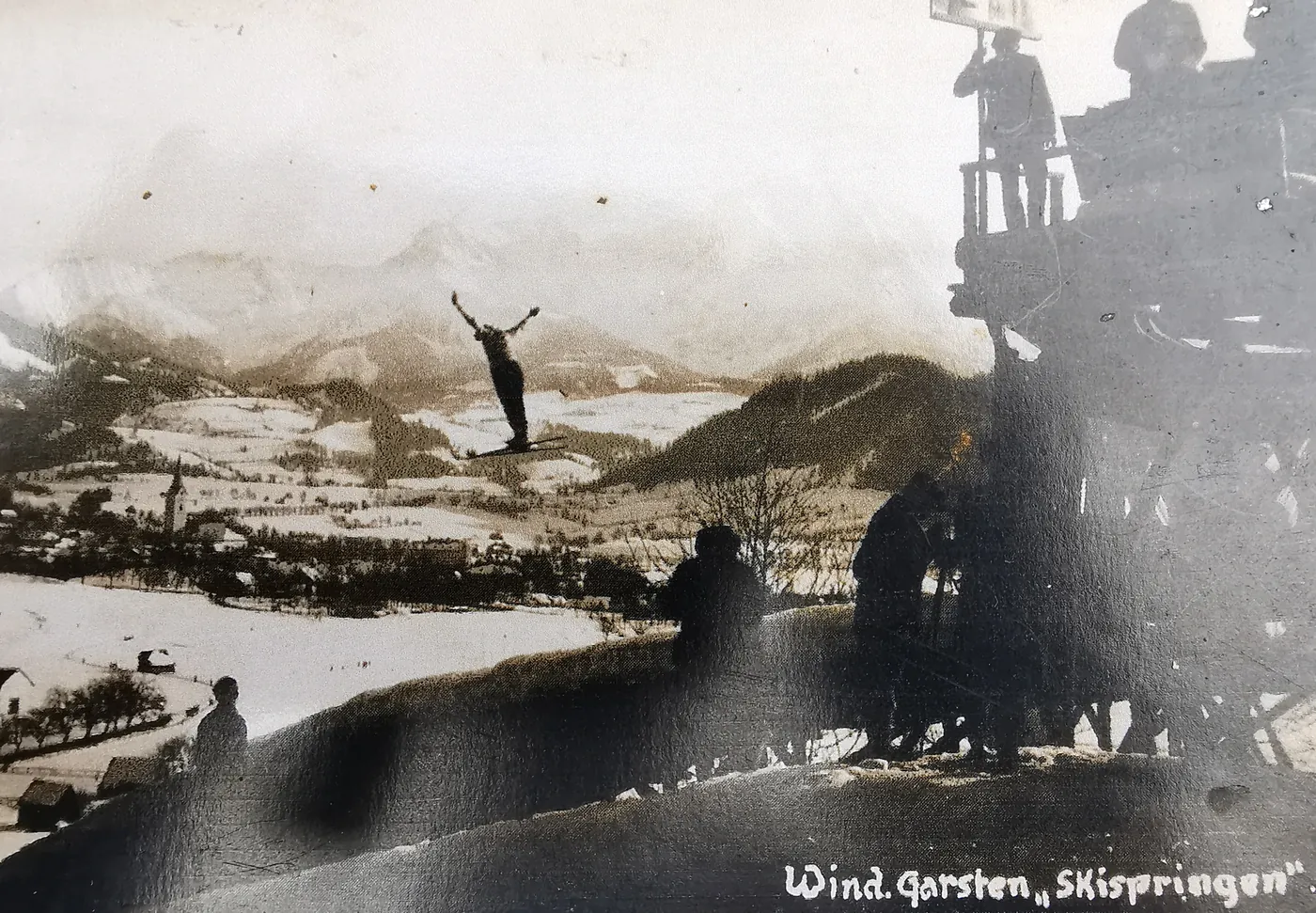 Ein Skispringer mit ausgebreiteten Armen in der Luft, unter ihm der verschneite Ort mit Kirchturm, rechts der Kampfrichterturm.